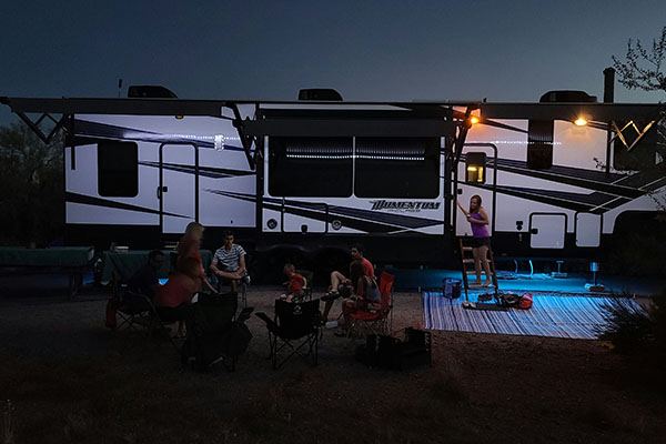 A family relaxes in camp chairs at dusk outside a Grand Design Momentum toy-hauler trailer lit by awning lights